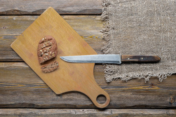 whole wheat bread with sunflower seeds and knife with cutting board and burlap on wooden background, top view 