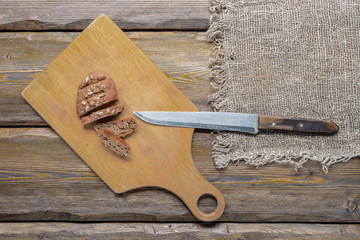 whole wheat bread with sunflower seeds and knife with cutting board and burlap on wooden background, top view 