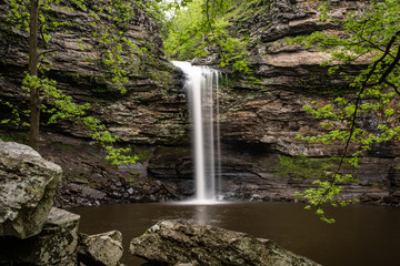 Fototapeta na wymiar Cedar Falls Petit Jean State Park Arkansas. The long waterfall fills the dark pool of water and dark rocks contrasted by green maple, pine and star shaped leaves of a sorghum tree frame it. 
