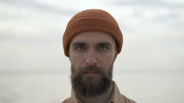 Portrait of no emotional, concentrated beard man looking at camera. Outdoors near the sea.