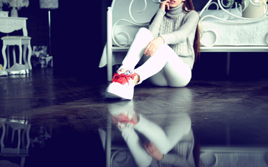 Forever young! Beautiful relaxed stylish girl sitting on a floor in white sneakers with red laces about a bed. reflection, posing at the camera against the grey background.
