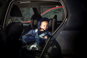 Unhappy, sad child sitting in a child car seat on the second row of a black car. He is dressed in warm overalls of dark blue color.