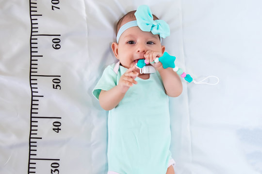 A Three Month Baby Girl In Mint Green Clothes Lying On A Bed On Which A Measuring Ruler For Growth Is Drawn. Teething Tool In Hands, Chewing