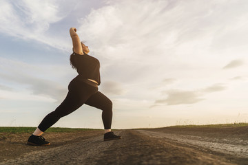 Young overweight woman doing morning yoga on the country road, open air activity. Healthy lifestyle, sport, weight loss - obrazy, fototapety, plakaty