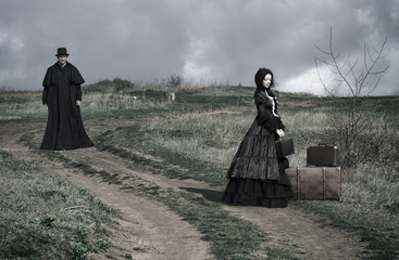 Outdoors portrait of a victorian lady in black sitting on the road with her luggage and gentleman...