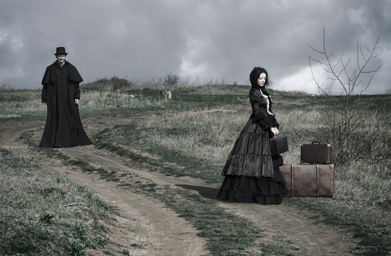 Outdoors portrait of a victorian lady in black sitting on the road with her luggage and gentleman walking down the road.