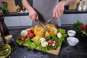 Cook holds knife and fork and chops chicken or turkey. Delicious food on wooden desk in kitchen. Grilled chicken or turkey with spices and fresh vegetables. Christmas or thanksgiving pay preparation