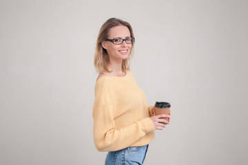 Fototapeta premium waist-up portrait of blonde woman holding coffee cap in her hands and smiling, looking at the camera. wearing yellow sweater and glasses. poses against grey background. face expression, emotions.