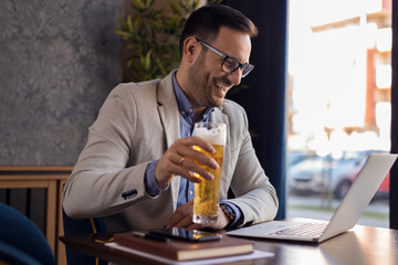 Mid adult smiling businessman drinking beer and surfing the net on laptop in a cafe