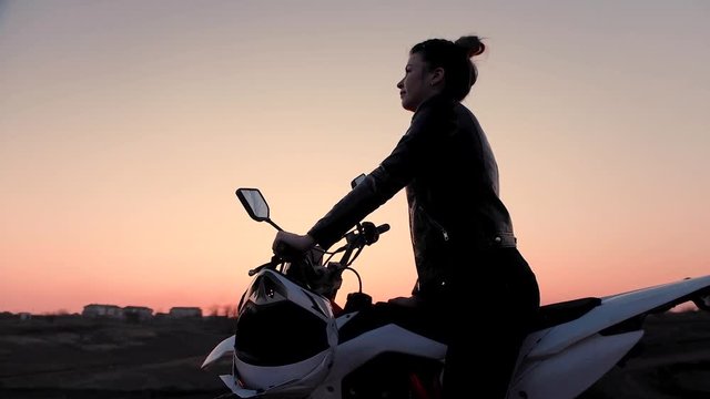 In the open, a young girl is sitting on a motorcycle watching a beautiful sunset