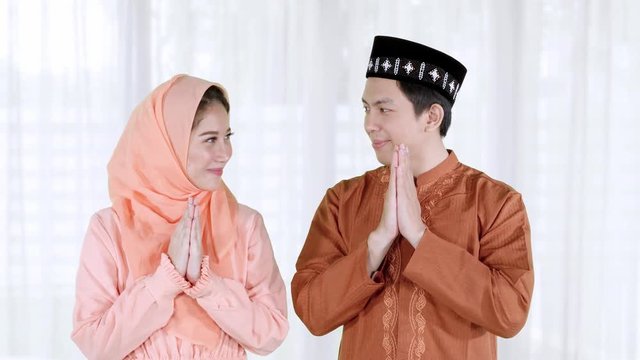 Two young attractive muslim couple showing a greeting hands gesture and smiling to each other while wearing islamic clothes during ramadan at home. Shot in 4k resolution