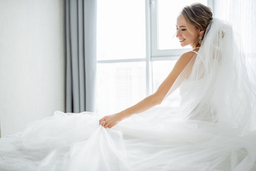 Amazing young bride posing in a beautiful wedding dress with wedding veil near window and smiling