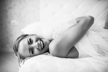 Black and white photo of a cheerful happy bride who is lying on the bed with wedding veil