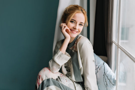 Portrait of smiling, happy blonde girl next to the window, relaxing in the morning, having good time at home. She's dressed in nice silk pijamas.