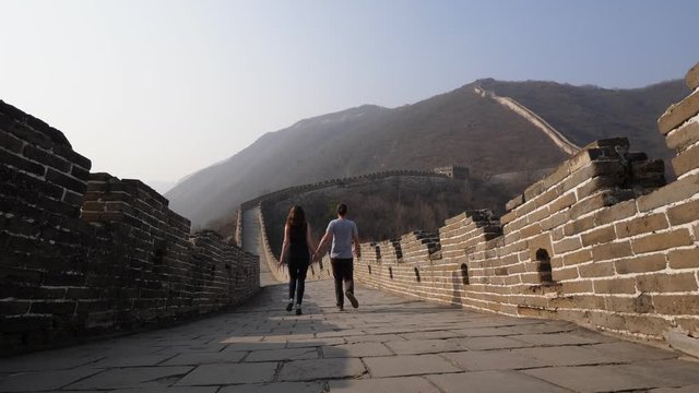 Young man and woman explore Great Wall of China together, low camera on stone pavement of wide passage. Tourists come down holding hands, enjoy empty Mutianyu site at evening time