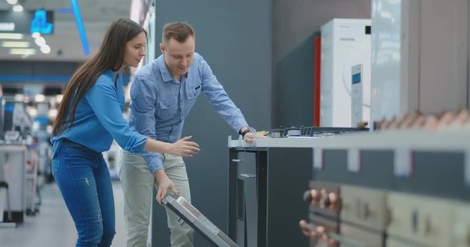 The man and the woman to open the door of dishwasher appliances in the store and compare with other models to buy the new house