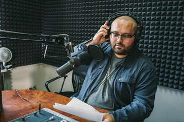 Man radio host or representer or journalist reads news from paper list in hand to studio microphone sitting at mixing sound panel or console at radio station
