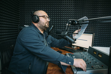 Man radio host or representer or journalist reads news from paper list in hand to studio microphone sitting at mixing sound panel or console at radio station