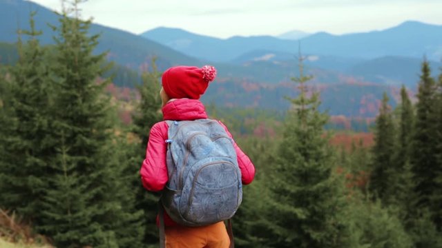 back view of a woman hiker with backpack, wearing in red jacket and orange pants, standing on the trail trail on the mountains and forest background, Western Ukraine.