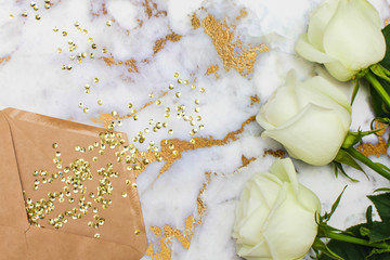 White roses and craft envelope lie on white and gold marble table. Top view, flat lay