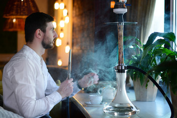 A bearded businessman in white shirt smokes a beige wooden hookah with lightning patterns in a...