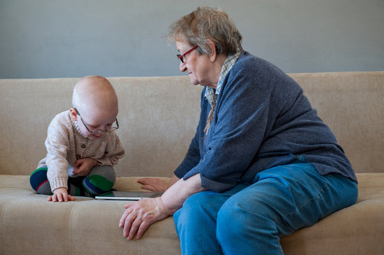 Elderly woman sits and plays with her grandson on couch. Child loves grandmother very much. Boy kisses and hugs his beloved grandmother. Kid shows how he can use computer tablet.