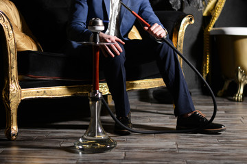 Fototapeta na wymiar Luxury businessman in a white shirt, blue jacket, dark pants and patent leather shoes sits on a black and gold armchair and smokes a red wooden hookah at home. The concept of rest.