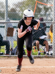 Obraz premium Female softball base runner smiling through her face mask while sprinting to first base.