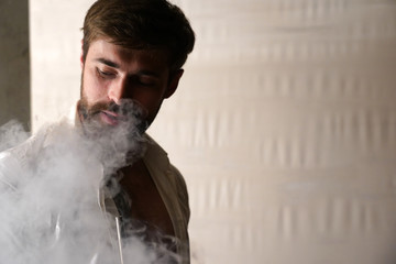 Portrait of a brutal bearded Caucasian man with short brown hair in a white shirt on blurred background. Handsome male smokes a hookah and exhales a cloud of smoke from his mouth with pleasure.