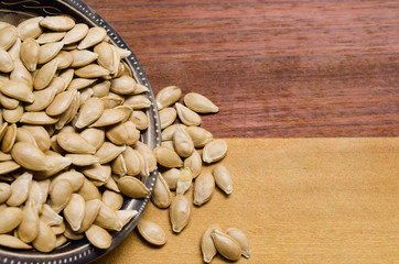 Pumpkin seeds inside a small bowl, with some falling seeds, on a two-colored wooden surface