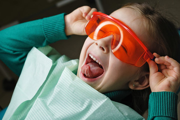Little girl sits in dental chair and wears protective glasses. She dabbles.