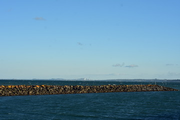Fototapeta premium Breakwall of free laid stones surrounded by dark choppy sea with Auckland cityscape in distant background.