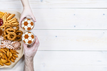 Hands holding glass mugs of beer  with a soccer ball on a beer foam on light wooden background. Empty space for text. Top view