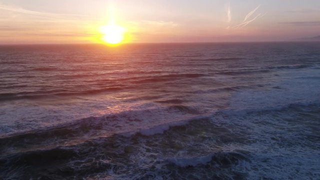 Drone captures a setting sun at golden hour over empty a rocky beach and surf break. Haze and setting sun create a dreamy and surreal vibe.