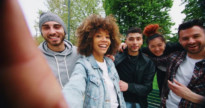 Slow motion of group of young friends of different ethnicities are having fun and making a selfie together with cellular phone in a green city park on a sunny day.