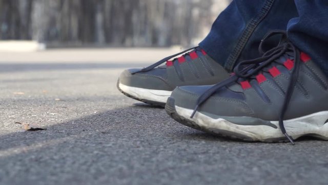 Close-up, feet of a man in sneakers and jeans sitting on a bench in a city park smoking a cigarette. Cigarette smoke and the falling cigarette butt which the person extinguishes, presses a foot