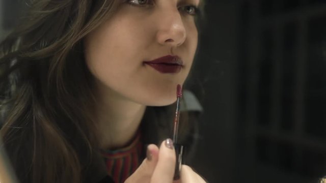 Brunette young woman putting dark red lipstick on lips while looking in mirror. Reflection of lady applying her make-up. Makeup at night getting ready before leaving