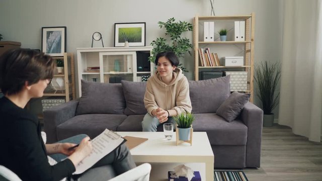 Zoom in of happy teenager cute boy talking to therapist smiling and laughing sitting on couch in office. Psychological help, positive emotions and childhood concept.