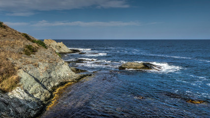 Fototapeta na wymiar Rocky cliff shore by a seaside, small waves, green horizon, blue sky with some smooth clouds