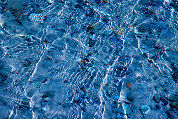 Blue water surface with ripples through which the sandy bottom with pebbles is visible
