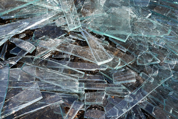 A pile of different sharp pieces of broken glass lying on the ground. Pieces of glass of different size and shape. Many pieces of oblong shape. The sharp edges glisten in the sunlight.
