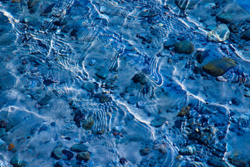 Blue water surface with ripples through which the sandy bottom with pebbles is visible