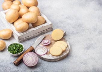 Fresh raw organic potatoes in wooden box and on chopping board with red and green onions on light table background.