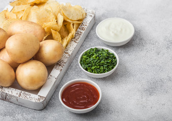 Fresh organic homemade potato crisps chips with raw yellow potatoes in white wooden box on light kitchen table background. Sour cream, ketchup and green onion