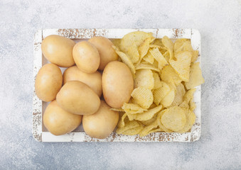 Fresh organic homemade potato crisps chips with raw yellow potatoes in white wooden box on light kitchen table background.