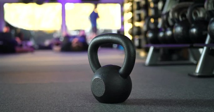 A kettlebell weight in a gym with people working out in a physical fitness training class in the background SLIDE LEFT.