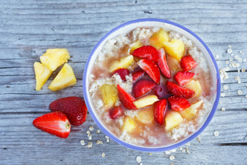 Oatmeal porridge in bowl topped with fresh strawberry and pineapple on old, rustic board. Top view.