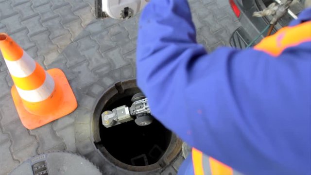 A worker is lowering specialized inspection camera equipment down a sewage drain.
