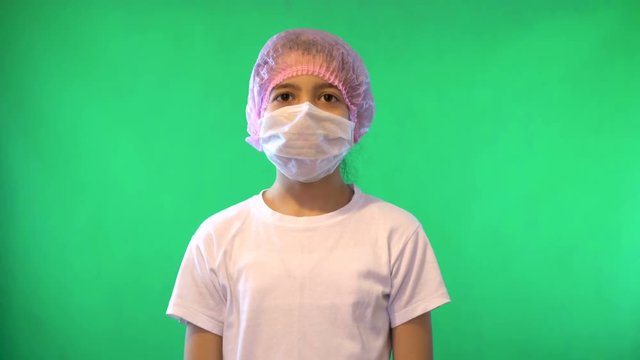 The girl stands on a green background and wears a medical mask on her fac