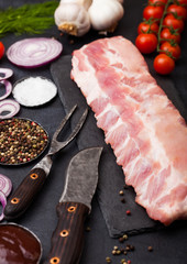 Fresh raw pork ribs on chopping board and vintage meat fork and knife on black stone background. Fresh tomatoes and red onion with garlic, salt and pepper.